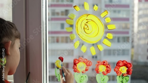 Little girl with brush in hand painting flowers and sun on the window at home. Colorful education at home, fun and play sensory 