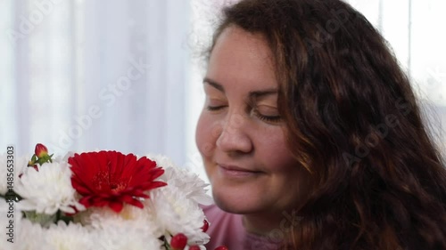 Brunett beautiful smiling woman breathing flavor of 
bouquet of flowers standing near the window at home