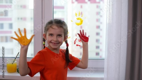 Little cute smiling girl leaves handprints in different colors of psintings on the window at home. Colorful education at home, fun and play sensory 