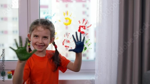 Little cute smiling girl leaves handprints in different colors of psintings on the window at home. Colorful education at home, fun and play sensory 