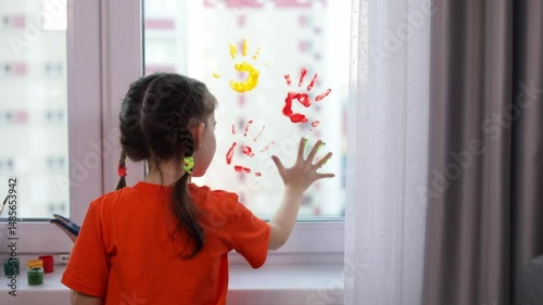Little cute smiling girl leaves handprints in different colors of psintings on the window at home. Colorful education at home, fun and play sensory 
