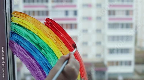 Woman's hand close up with paint brush painting colorful Rainbow on the window at home 