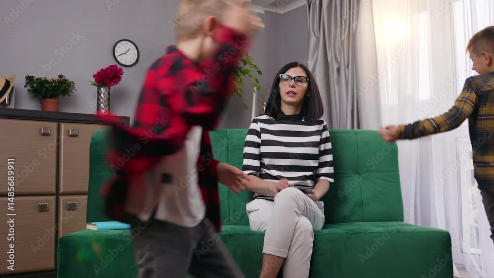 Tired young woman in glasses working with tablet pc while two noisy loud sons interfering her and running around sofa on which she sits