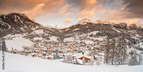 Moena village, Fassa Valley, Trentino, Italy. Snow-covered houses nestle in a valley surrounded by mountains, with a cloudy sky tinged with orange at sunrise.