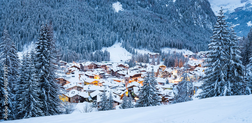 Canazei Village, Sella Pass, Fassa Valley, Trentino, Italy. Snow-covered houses and trees are illuminated at dusk, nestled against a backdrop of a dense, snow-laden forest.