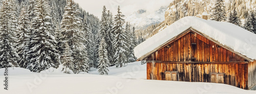 A snow-covered wooden cabin stands in a winter landscape, surrounded by snow-laden pine trees and mountains. San Nicolò Valley, Fassa Valley, Trentino, Italy. 