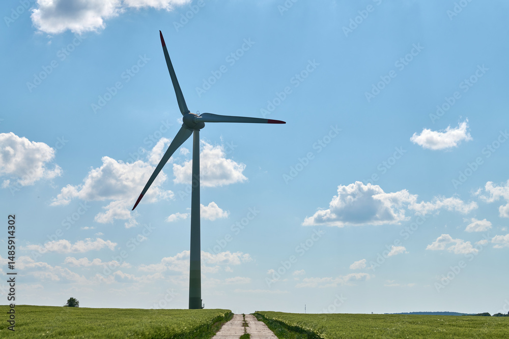 Obraz premium Wind turbine with red tipped blades stands beside cultivated wheat farmland and straight concrete road under clear blue sky evoking balance between human effort and natural resources