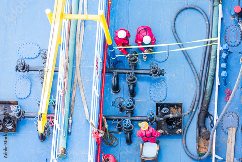 Top-down view of a bunker barge moored alongside a large merchant vessel. Crew in red coveralls and white helmets are busy on deck, captured during routine fuel transfer operations.
