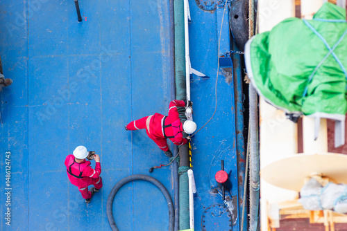 Top-down view of a bunker barge moored alongside a large merchant vessel. Crew in red coveralls and white helmets are busy on deck, captured during routine fuel transfer operations.