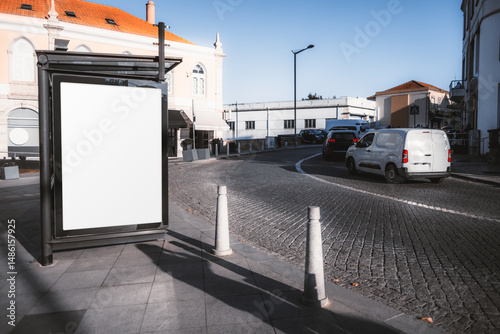 Wallpaper Mural A blank, vertical mockup billboard on a city sidewalk, on the bus stop, next to a cobblestone road, with cars driving on a curved street and historic European-style buildings in the background Torontodigital.ca