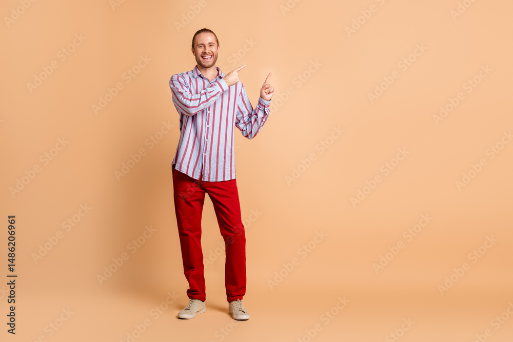 Young happy man in casual striped shirt making pointing gesture standing against beige background promoting advertisement Young happy man in casual striped shirt making pointing gesture standing against beige background promoting advertisement
