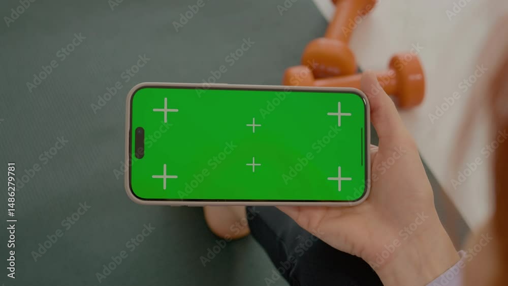 A woman sits on a gym mat in her living room, looking at a smartphone with a green screen while taking a break from her workout routine.
