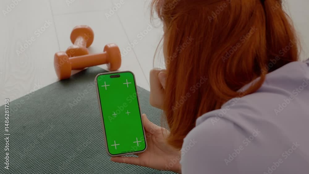 A woman with red hair rests on a gym mat, holding a green screen smartphone. She appears focused as she examines her device during a break from exercising.