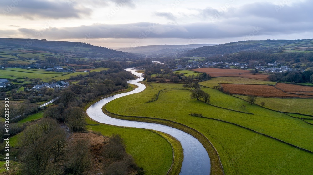 Fototapeta premium Aerial view captures the graceful curve of a river meandering through vibrant farmland and quaint towns, framed by lush hills and forests under a cloudy sky