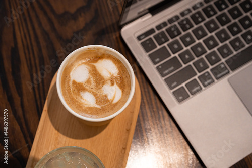 Close-up of Hot coffee latte with latte art milk foam in cup made of paper and laptop computer on wood desk office desk in coffee shop at the cafe,during business work concept
