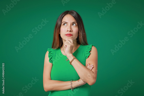Young Latina woman with black hair and green shirt in expression of thought and doubt, on a vibrant green background