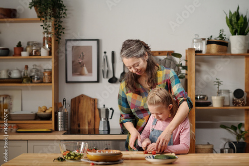 Wallpaper Mural Mother and daughter preparing meal in cozy kitchen with wooden shelves and green plants. Mother embracing daughter while assisting in slicing vegetables on cutting board Torontodigital.ca
