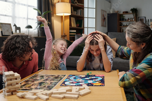 Family gathered around table, celebrating a win during competitive board game night, with expressions of joy and camaraderie evident. Homey background filled with cozy details