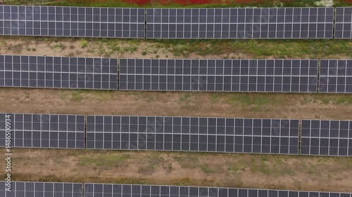 Drone shot of large-scale photovoltaic solar panel installation in rural farmland surrounded by lush greenery and cloudy weather
