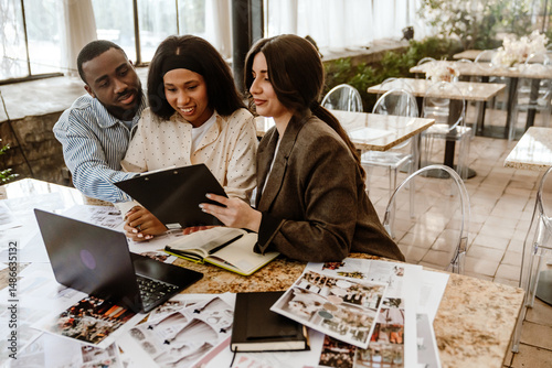 A young Black bride and a young Black groom are sitting beside a young White female wedding planner in a restaurant, reviewing venue photographs and notes for their upcoming wedding celebration.