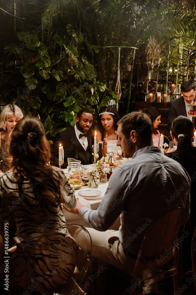 Obraz premium The wedding guests enjoy a candlelit dinner at an elegant indoor reception as the bride and groom sit among them, surrounded by lush greenery and ambient lighting.