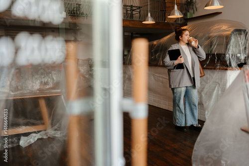 A young White female cafe owner in a gray oversized blazer and blue jeans sipping coffee while holding a clipboard inside her under-renovation cafe with covered furniture and an unfinished interior.