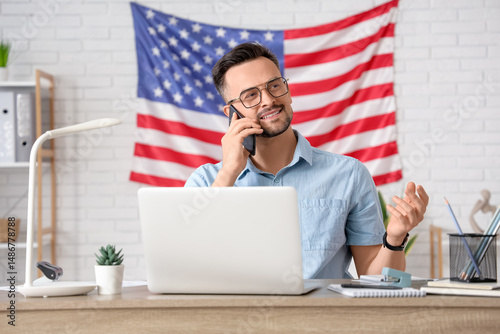 Handsome young happy man with laptop and flag of USA talking by mobile phone ...