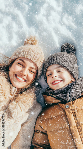 Mother and son smiling while lying in the snow, dressed in winter clothes and enjoying a joyful moment as snow falls.