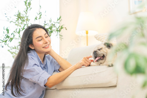 Happy Asian woman enjoy and fun playing with senior Pug dog breed in living room. Beautiful woman relaxing with domestic dog at home. Pet humanization, pet parent and senior pet healthcare concept.