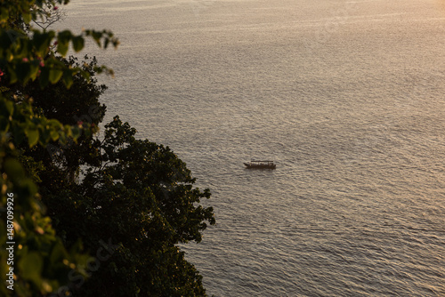 Traditional wooden boat floating on calm sea at sunset, viewed from the tropical cliffs of Uluwatu temple in Bali. Trees and plants in the foreground, ocean in the background. Golden hour in Indonesia