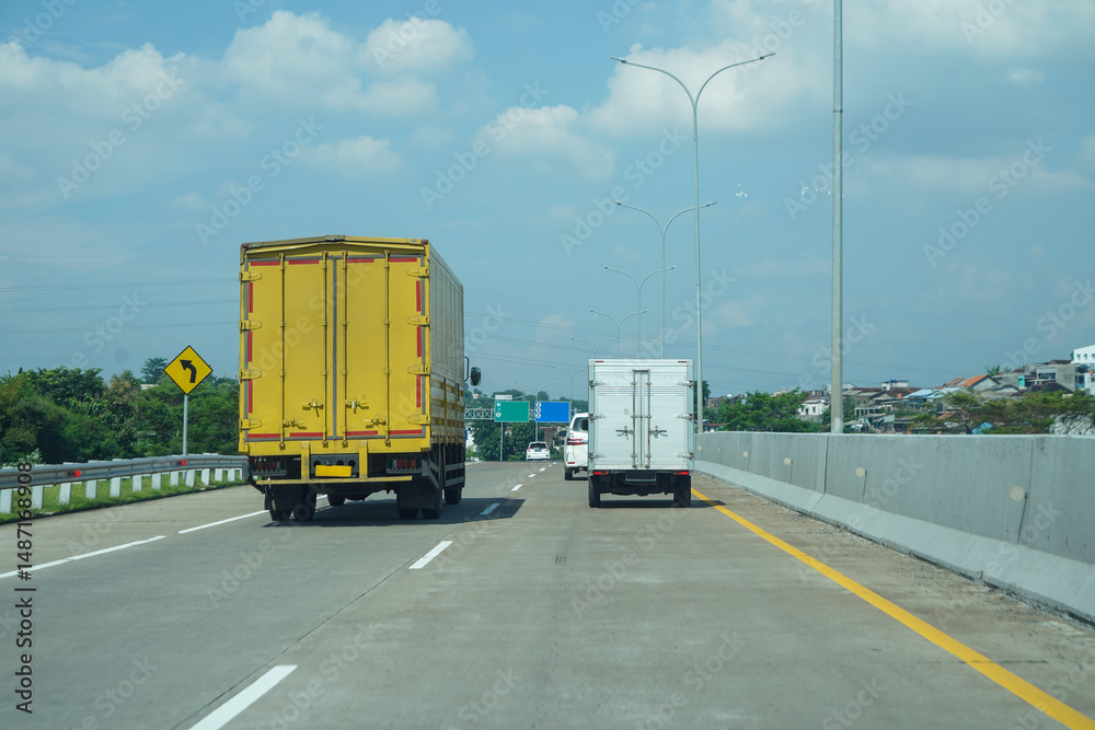 Naklejka premium Trucks Driving on Highway Under Blue Sky with Urban Background and Traffic Sign