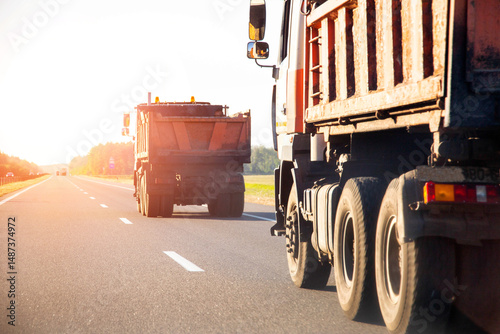 Orange road dump trucks drive along the highway after delivering asphalt against the backdrop of sunset in summer, road repair. Copy space for text, industry