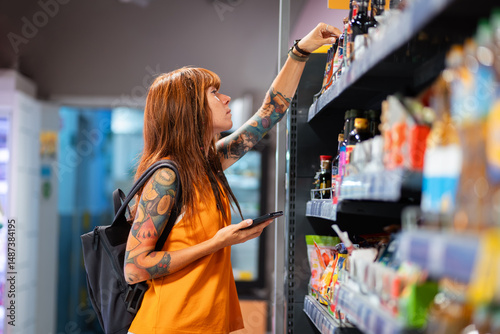 Side view of young pretty tanned Caucasian woman with tattoo reaches hand to top shelf in supermarket. Shopping and consumerism