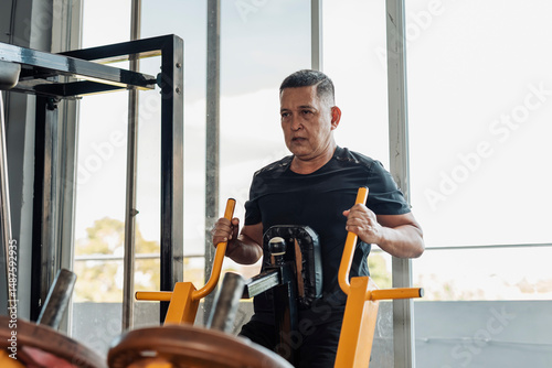 A determined Southeast Asian man in his 50s performs seated rows on a plate-loaded chest supported row machine at an open-air gym. Training back.
