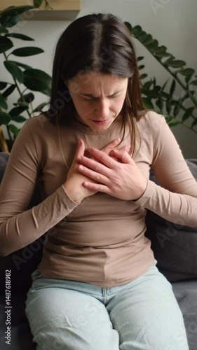 Vertical video of unhealthy young caucasian woman having severe chest pain while sitting alone on sofa, sad worried female suffers from heartache at home, infarction or heart disease concept