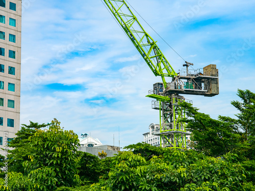 A tall green construction crane towers above city buildings and lush trees under a blue sky, illustrating urban development and architectural progress in a growing city.