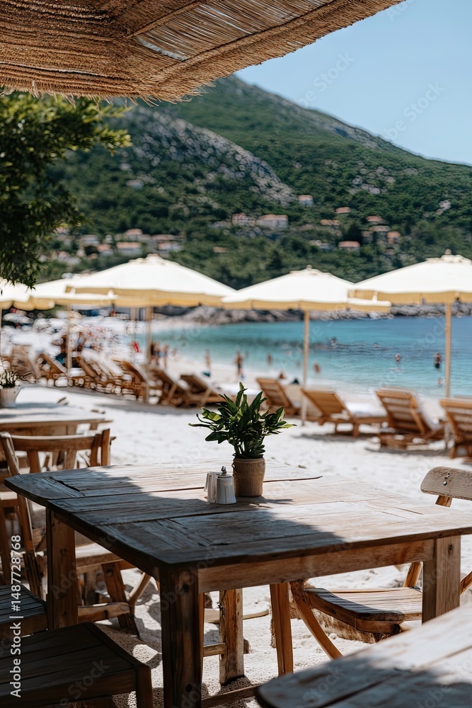 Fototapeta premium A serene beachside dining setup with wooden tables and chairs under large umbrellas. overlooking a calm sea and lush green hills in the background. ideal for relaxation and leisure