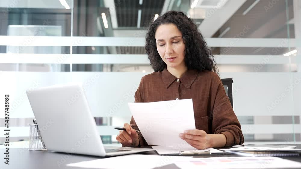Happy businesswoman satisfied with financial results sitting at workplace in business office. Smiling female financier looks at documents and computer screen, rejoices at positive indicators, success