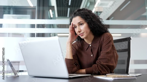 Upset shocked businesswoman reads bad news on a laptop while sitting at a desk at workplace in a modern business office. Frustrated worried woman is disappointed by a negative message on the computer