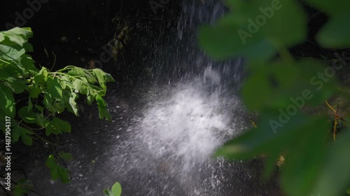 Right-to-left pan reveals a waterfall behind vibrant green foliage as sunlight sparkles on splashing water in a serene forest scene