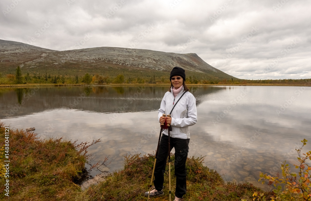 Fototapeta premium Woman hiking in Swedish Lapland during autumn, standing by a tranquil mountain lake with Nordic hills in the background. Scenic wilderness adventure in Scandinavia with vibrant fall colors