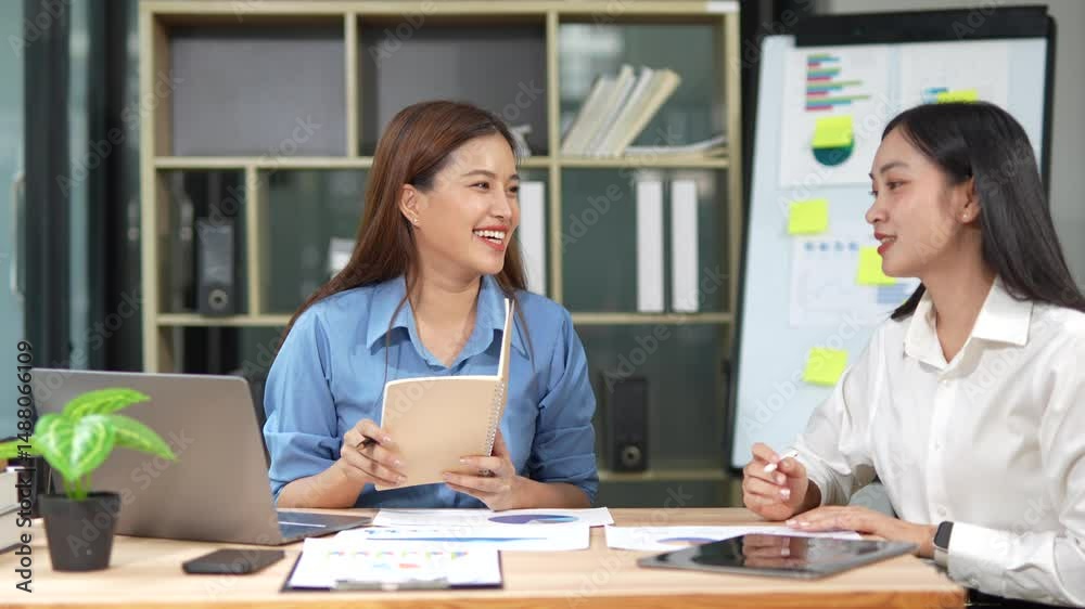 Two Asian businesswomen discussing a project on a laptop sitting at a desk in an office. Business people working together. Two Asian businesswomen discussing a project on a laptop sitting at a desk in an office. Business people working together.