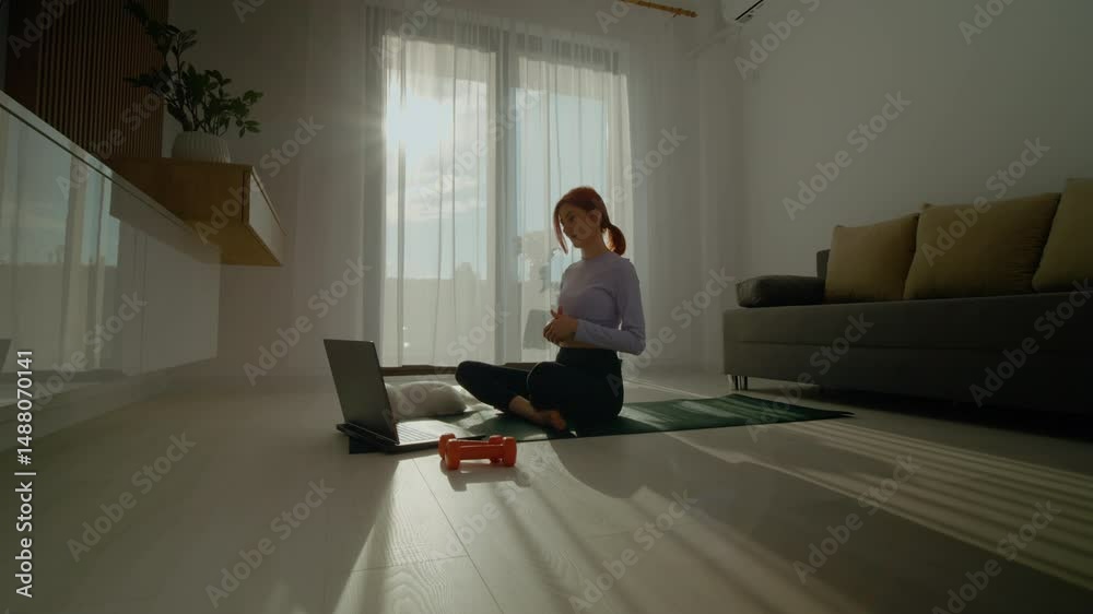 A redhead woman sits on a gym mat in her living room, engaging in an online fitness class with her laptop on a sunny morning while surrounded by dumbbells and her cat.