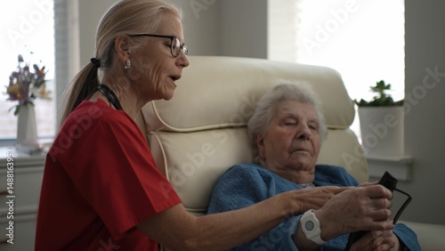 A caregiver helps an elderly woman with a mobile device in a bright, comfortable living room during daylight hours, showcasing a moment of connection.