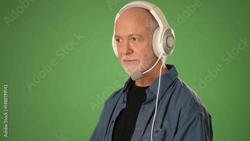 A mature man wearing a customer service headset isolated against a solid green screen, expressing emotion while talking to customer.