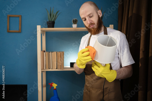 Bearded man washing and cleaning lamp with rag from dust in living room