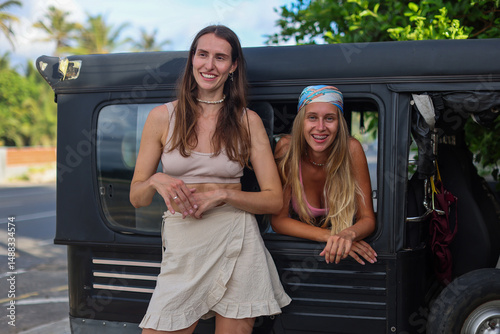 Two joyous women are enjoying life together beside a vintage jeep in a beautiful tropical paradise