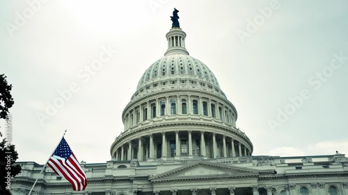 The US Capitol building dome stands majestically against a cloudy sky with an American flag waving gently in the foreground. The iconic white dome remains stationary as the flag flutters in the breeze