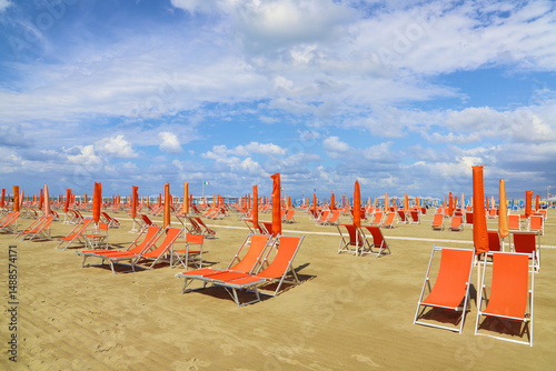 Wallpaper Mural Rows of orange beach chairs and closed umbrellas are neatly arranged on a sandy beach under a partly cloudy blue sky. The chairs, with matching sun umbrellas, create a vibrant, orderly scene. Torontodigital.ca