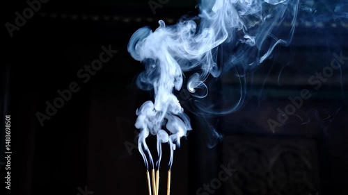 Mystical aroma of burning incense sticks releasing swirling wisps of smoke against a dark backdrop creating abstract shapes and patterns
